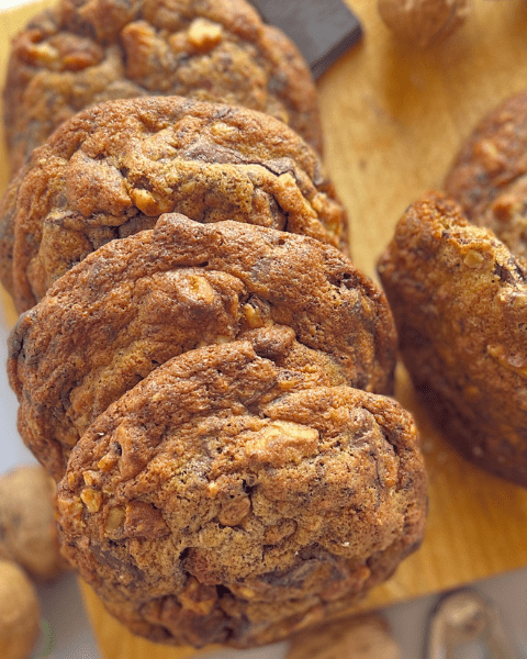 Maxi cookies dorés à point, croquants et moelleux à la fois, aux noix torréfiées et morceaux de chocolat noir présentés sur une planche en bois, avec des noix en arrière-plan.