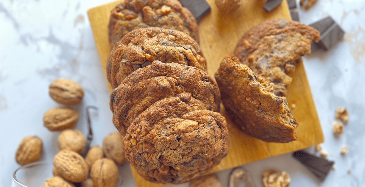 Maxi cookies dorés à point, croquants et moelleux à la fois, aux noix torréfiées et morceaux de chocolat noir présentés sur une planche en bois, avec des noix en arrière-plan.