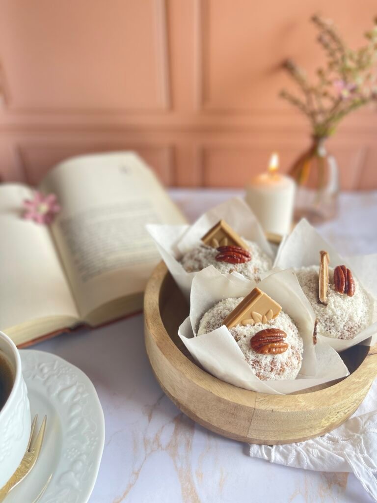 Muffins Dulcey coco et noix de pécan présentés dans des caissettes en papier, décorés de chocolat blond et de noix de pécan, accompagnés d'un café dans un cadre cosy. Recette sur Deliciously Home.