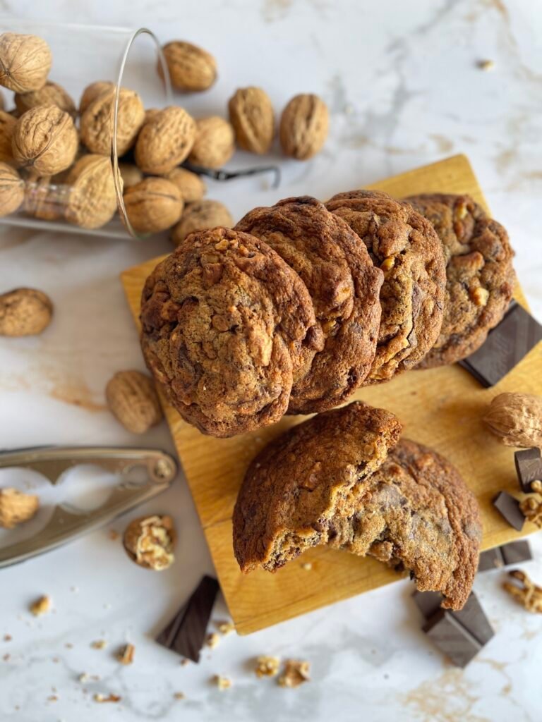 Vue plongeante de maxi cookies chocolat noir et noix torréfiées sur une planche, entourés de noix et de morceaux de chocolat, texture dorée et croustillante visible.