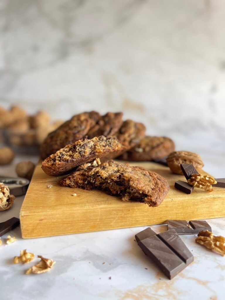 Vue d'une de maxi cookies chocolat noir et noix torréfiées sur une planche, entourés de noix et de morceaux de chocolat, texture dorée et croustillante visible.