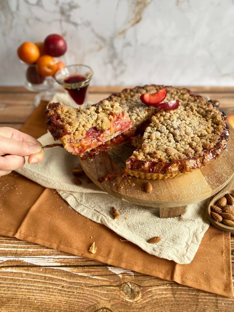 Une main servant une part de tarte crumble prunes & porto avec une croûte croustillante, accompagnée d'un verre de porto. Recette disponible sur Deliciously Home.