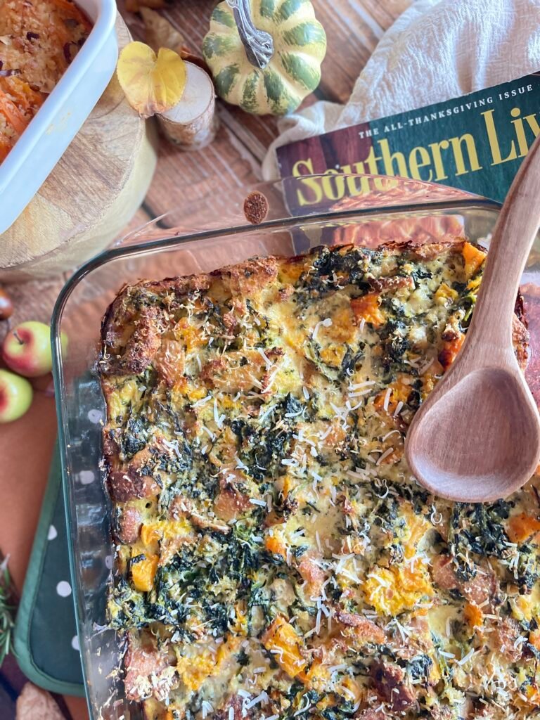 Un grand plat en verre contenant un pudding au pain salé à la courge, aux épinards et au parmesan râpé, posé sur une table décorée de citrouilles, pommes et un magazine de Thanksgiving.