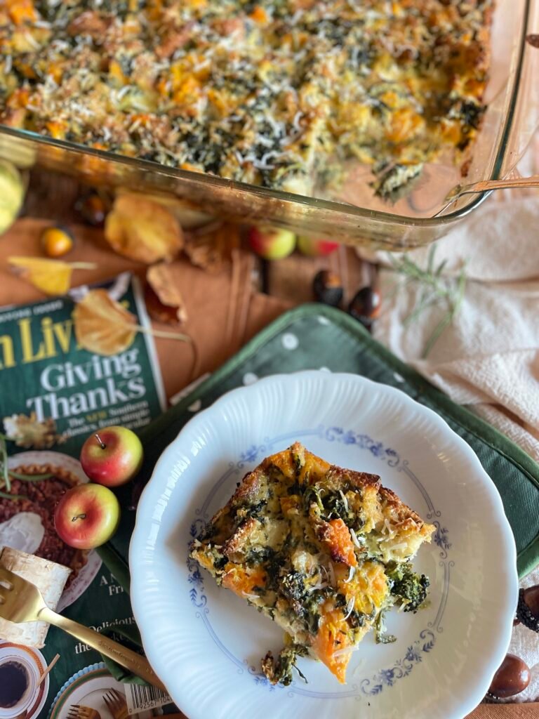 Une assiette en porcelaine blanche et bleue contenant une portion de pudding salé à la courge et aux épinards, accompagnée d'une décoration de pommes miniatures et d'un magazine de cuisine en arrière-plan.