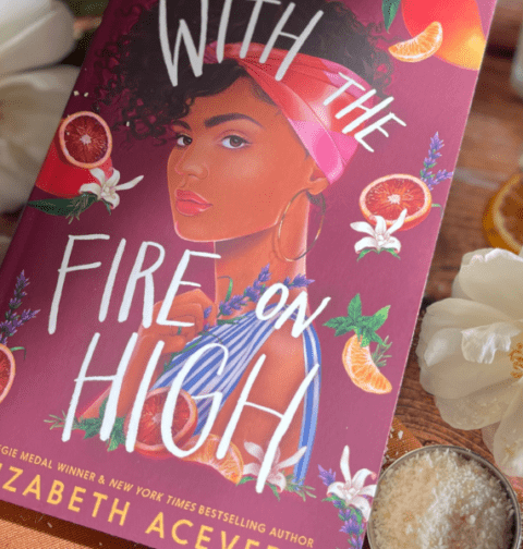 Le livre "With the Fire on High" d'Elizabeth Acevedo posé sur une table en bois, entouré de fleurs blanches, de tranches d'orange séchées et de romarin. Retrouvez notre Book Club sur Deliciously Home.