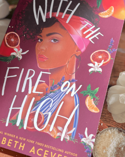 Le livre "With the Fire on High" d'Elizabeth Acevedo posé sur une table en bois, entouré de fleurs blanches, de tranches d'orange séchées et de romarin. Retrouvez notre Book Club sur Deliciously Home.