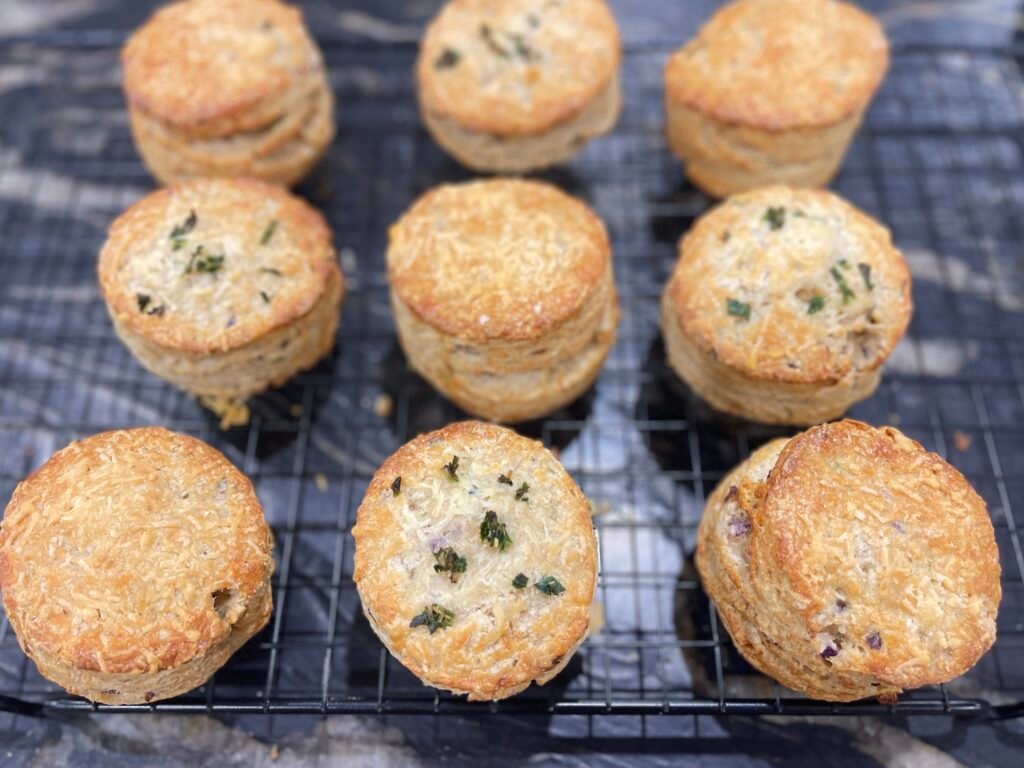 Disposition de scones dorés et uniformément espacés sur une grille de refroidissement, garnis de ciboulette fraîche et parsemés de parmesan.