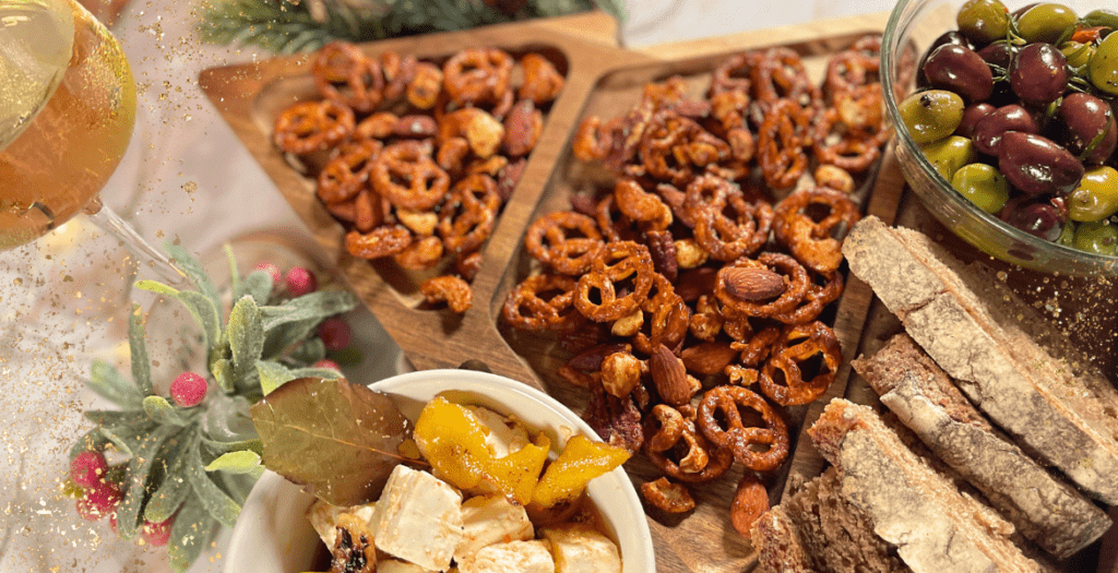 Un gros plan sur une table d’apéritif avec des bols d’olives, des bretzels épicés, des amandes et une verrine de feta marinée, entourés d’éléments décoratifs hivernaux et festifs. Recette sur Deliciously Home.