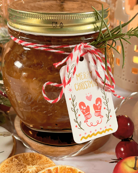 Un pot de chutney maison fini, décoré pour Noël avec une étiquette festive et entouré de tranches d'orange séchées, de pommes miniatures et d'éléments décoratifs de Noël, sur une table blanche lumineuse. Recette sur Deliciously Home.