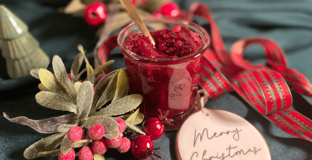 Vue rapprochée d’un pot de sauce aux airelles, sa texture rubis mise en valeur avec des décorations festives. Découvrez la recette sur www.deliciouslyhome.com.
