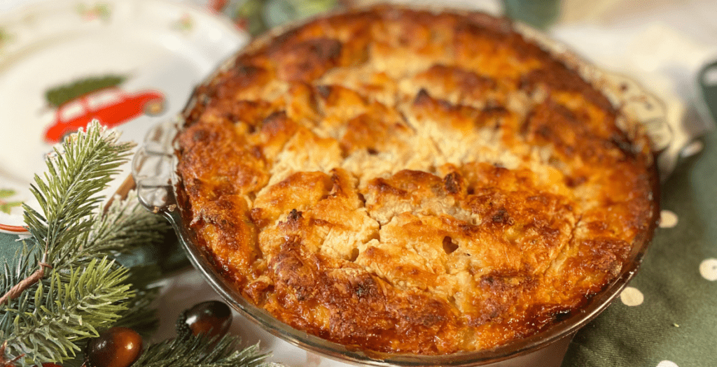 Vue rapprochée d'un plat de strata dorée et moelleuse, posée sur une table de fêtes, décorée de branches de sapin et d'ornements de Noël. Recette sur Deliciously Home.