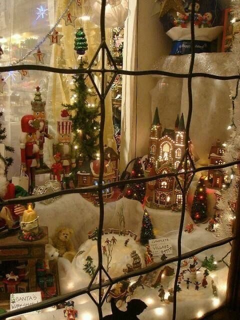 Village miniature de Noël vu à travers une vitrine décorée, avec des maisons illuminées, des sapins, des figurines et des ornements rétro. Une scène captivante illustrant l'esthétique Nostalgia-Core et la magie des fêtes d'antan. Article sur Deliciously Home.