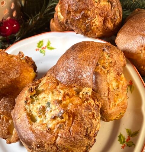 Ensemble de popovers dorés et moelleux placés sur une assiette décorée, avec des décorations de Noël en arrière-plan, incluant un petit sapin et des ornements festifs. Recette sur Deliciously Home.