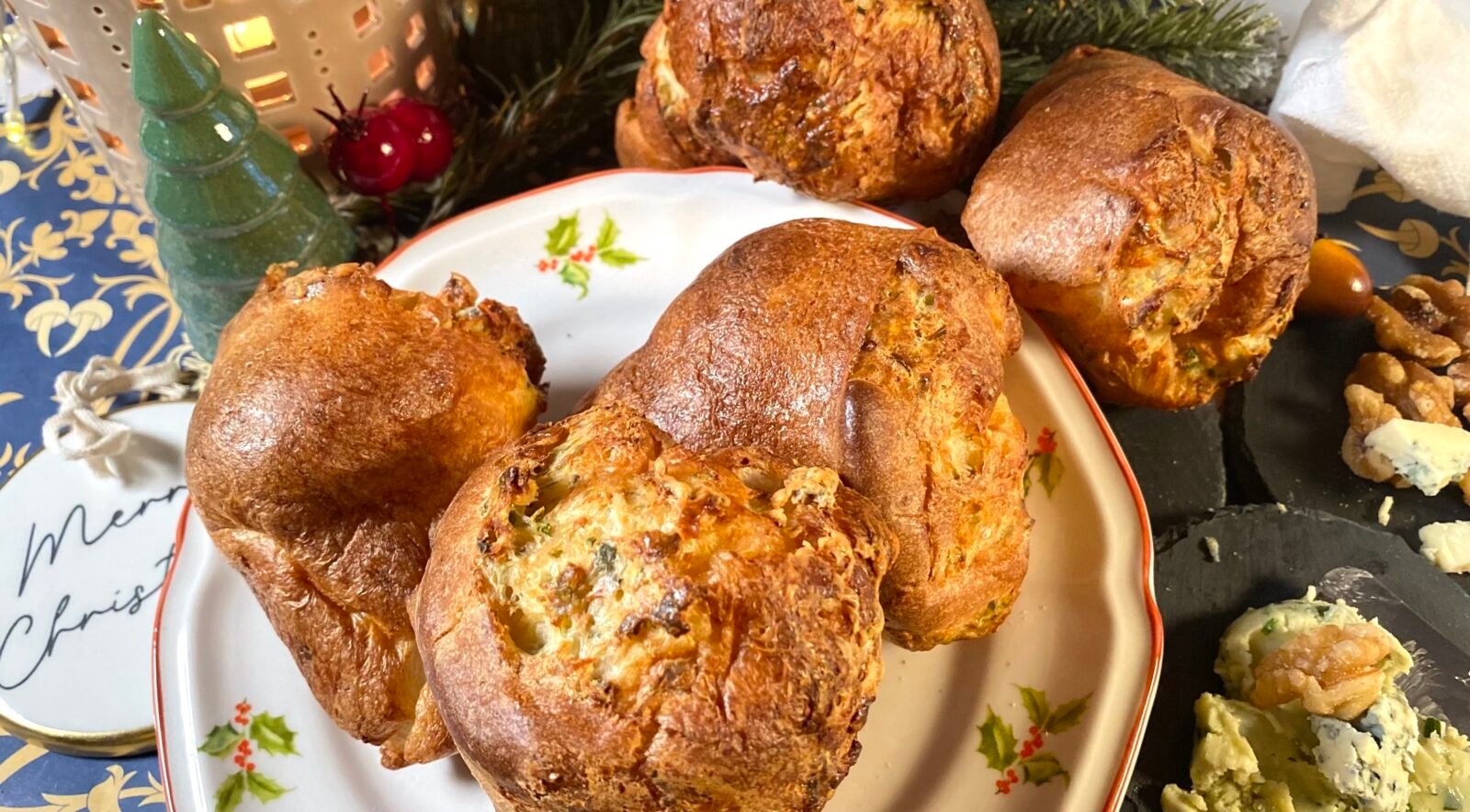 Ensemble de popovers dorés et moelleux placés sur une assiette décorée, avec des décorations de Noël en arrière-plan, incluant un petit sapin et des ornements festifs. Recette sur Deliciously Home.