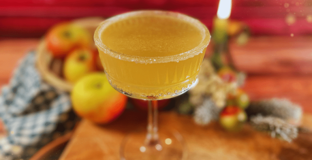 Vue large d'un verre de cocktail doré posé sur une table en bois, entouré de pommes et de décorations hivernales, avec une lumière tamisée en arrière-plan. Retrouvez la recette sur www.deliciouslyhome.com