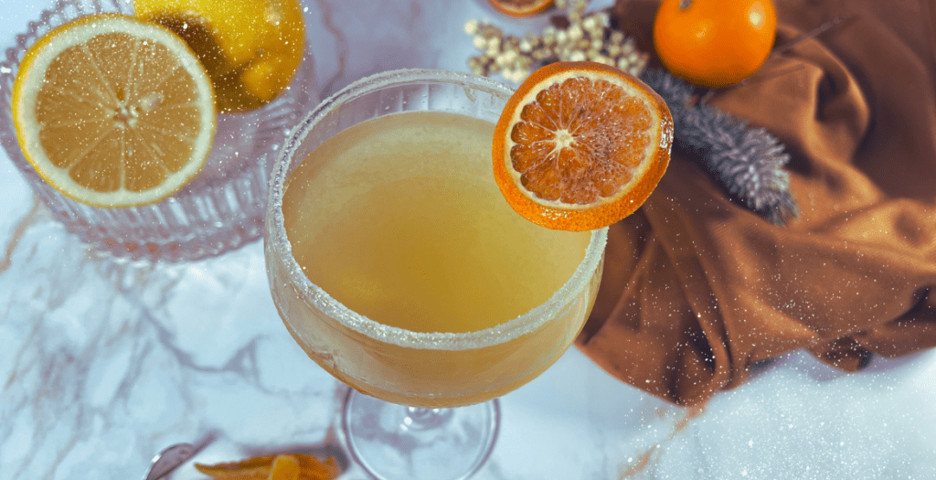 Vue aérienne d’un cocktail doré dans un verre à pied, orné d’une tranche d’orange séchée, avec des agrumes frais et des décorations hivernales en arrière-plan. Retrouvez la recette sur www.deliciouslyhome.com