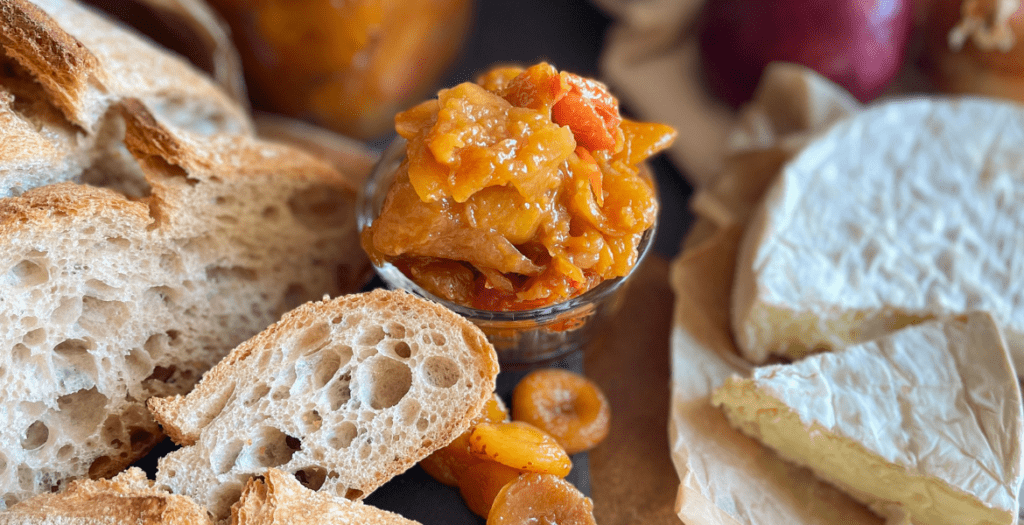 Une cuillère de chutney d’abricots et tomates maison, accompagné de pain rustique et fromage fondant, pour une expérience gustative équilibrée entre douceur et acidité. Recette disponible sur www.deliciouslyhome.com.