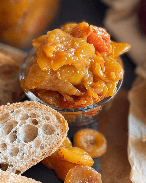 Une cuillère de chutney d’abricots et tomates maison, accompagné de pain rustique et fromage fondant, pour une expérience gustative équilibrée entre douceur et acidité. Recette disponible sur www.deliciouslyhome.com.