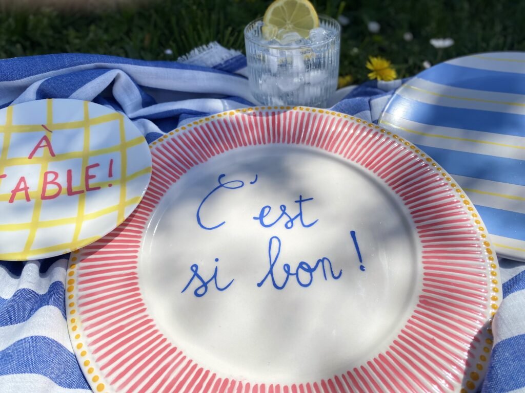 rayures assiettes printemps deliciouslyhome article4 Table printanière avec vaisselle colorée et message 'À TABLE !' et 'C'est si bon !', illustrant les Happy Hours Créatifs de Deliciously Home dans un décor ensoleillé et convivial.