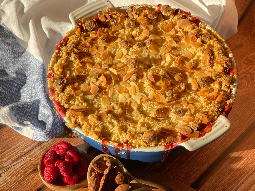 crumble rhubarbe framboise marzipan article1 llustration du crumble d’automne rhubarbe-framboise-marzipan fraîchement sorti du four, avec ses amandes effilées dorées et son jus légèrement caramélisé : un visuel évocateur de la chaleur réconfortante de cette recette signée Deliciously Home.