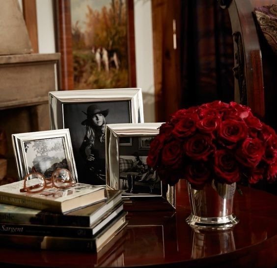 Table d’appoint avec bouquets de roses rouges, cadres photo argentés, livres et lunettes — évocation du style Ralph Lauren entre tradition, héritage et chaleur familiale.