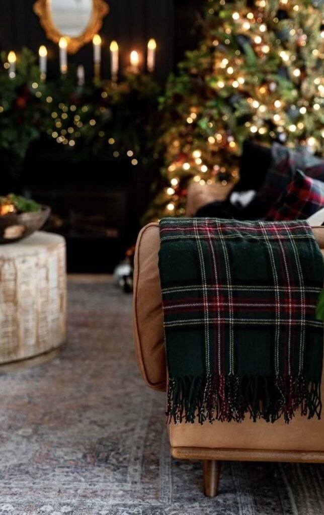 Plaid tartan vert foncé et rouge posé sur un canapé camel, avec en arrière-plan un sapin de Noël illuminé et une cheminée décorée.
