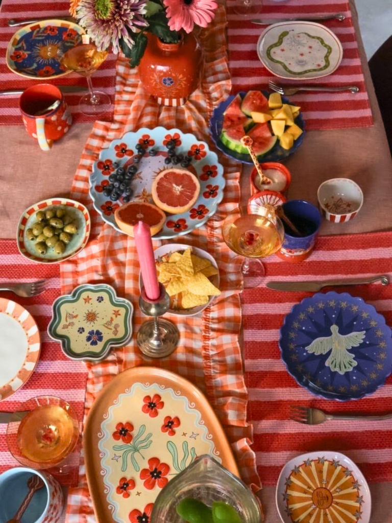 Grande table dressée avec une nappe rayée orange et rose, de la vaisselle aux motifs vibrants, des fleurs fraîches et des aliments colorés, créant une ambiance festive à découvrir sur Deliciously Home.