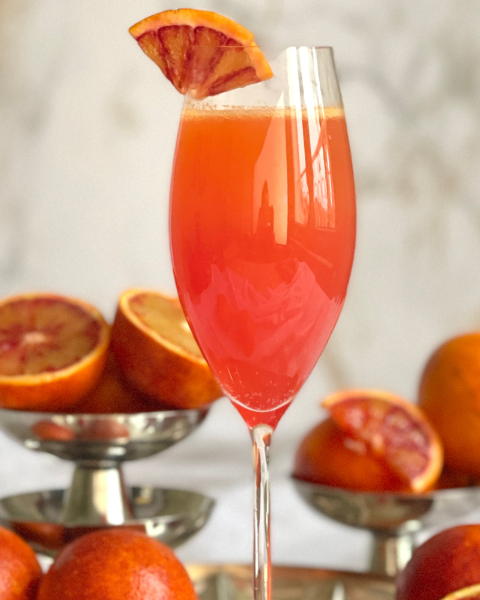 Flûte remplie d’un mimosa orange sanguine décorée d’une tranche d’orange sanguine, posée devant des oranges sanguines entières et coupées, cocktail à découvrir sur Deliciously Home.