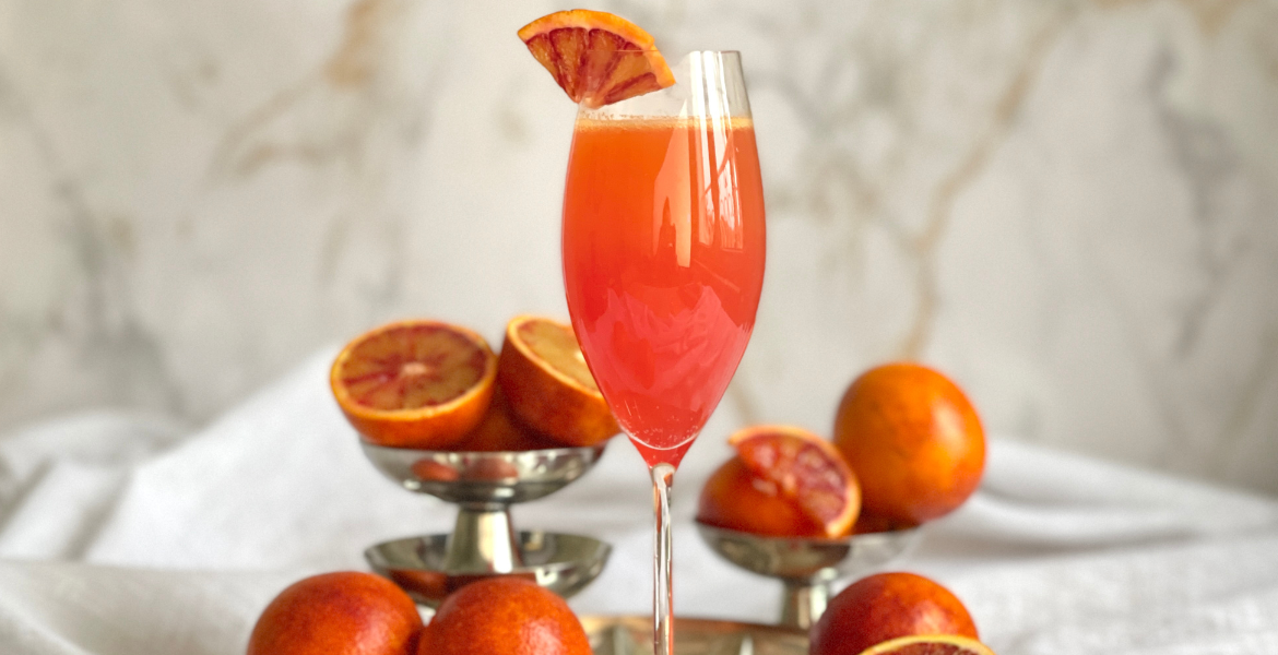 Flûte remplie d’un mimosa orange sanguine décorée d’une tranche d’orange sanguine, posée devant des oranges sanguines entières et coupées, cocktail à découvrir sur Deliciously Home.