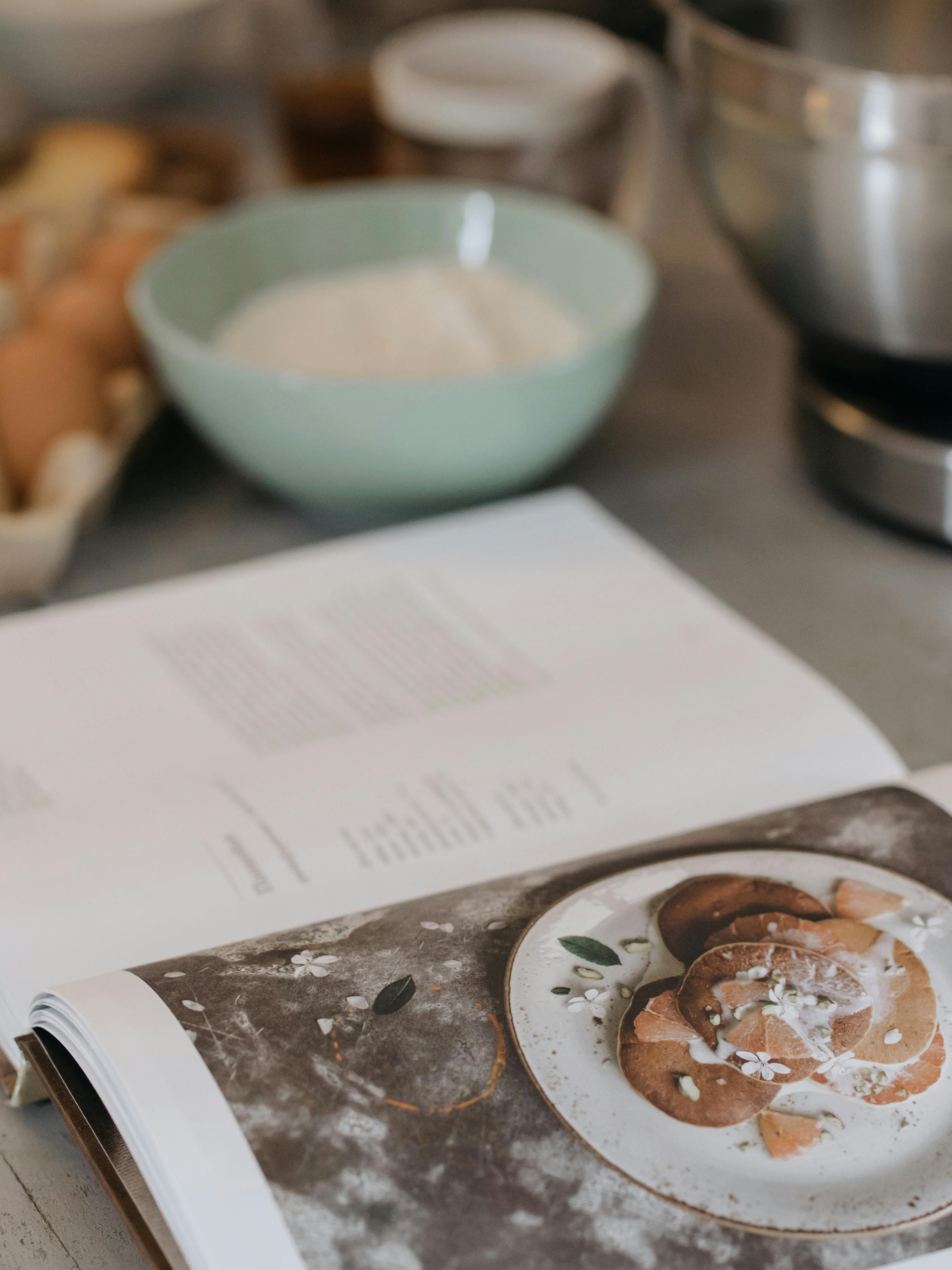 Livre de cuisine ouvert sur une recette, posé à côté d’ingrédients et d’un bol de farine dans une ambiance chaleureuse. Découvrez nos idées lecture sur Deliciously Home.