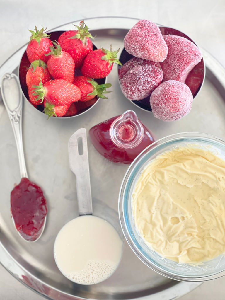 Ingrédients pour milkshake fraise vanille : fraises fraîches et surgelées, glace vanille, lait, confiture et sirop de fraise sur un plateau