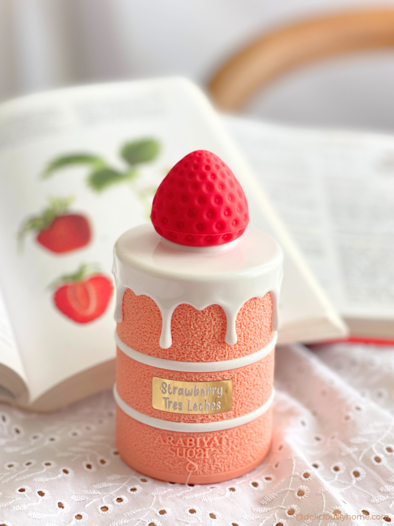 Strawberry Tres Leches d’Arabiyat Sugar posé devant un livre de cuisine illustré de fraises, en lien avec notre signature parfumée d’avril sur DeliciouslyHome, où nous partageons notre avis ainsi que les notes olfactives du parfum, entre fraise fraîche, lait et touche citronnée.