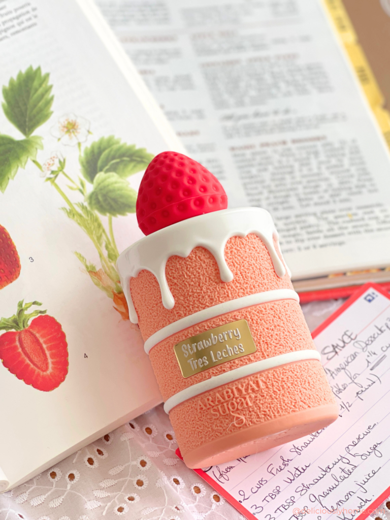 Le flacon de Strawberry Tres Leches d’Arabiyat Sugar photographié sur des livres de recettes et une fiche cuisine, illustrant l’univers de l’article DeliciouslyHome, où nous détaillons notre avis et les notes du parfum, mêlant fraise, accords lactés et douceur sucrée.