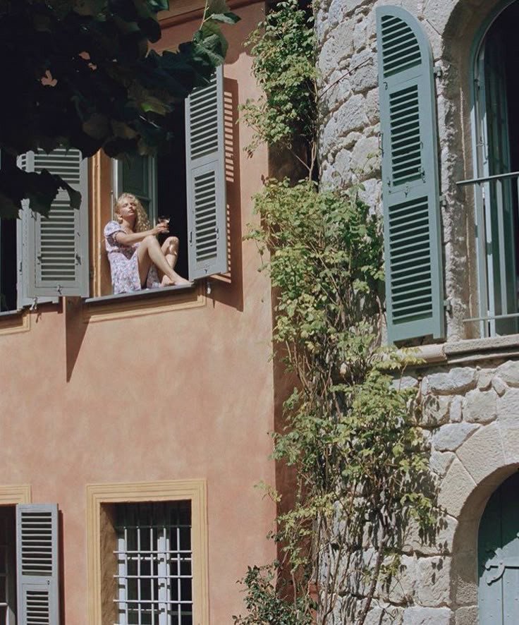 Femme assise à la fenêtre ouverte profitant de l’air d’été dans une ambiance méditerranéenne, à retrouver dans notre article sur Aménager un coin d’été chez soi sur Deliciously Home