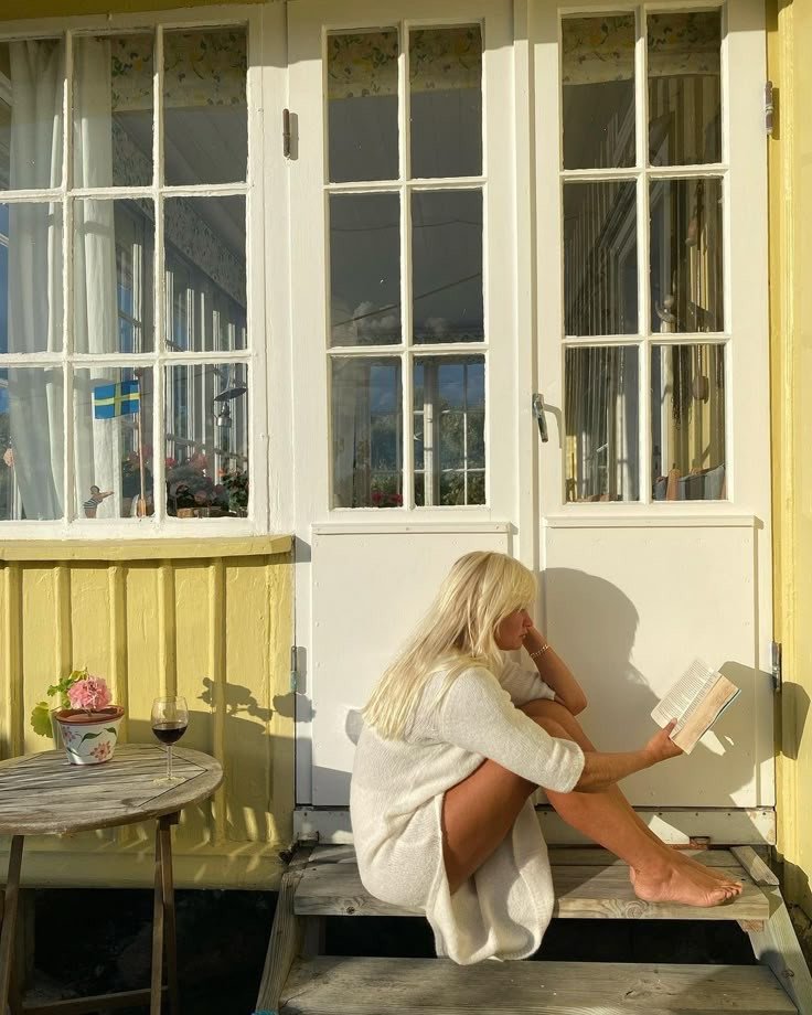 Femme assise sur des marches en bois lisant au soleil devant une maison jaune, à découvrir dans notre article sur Aménager un coin d’été chez soi sur Deliciously Home