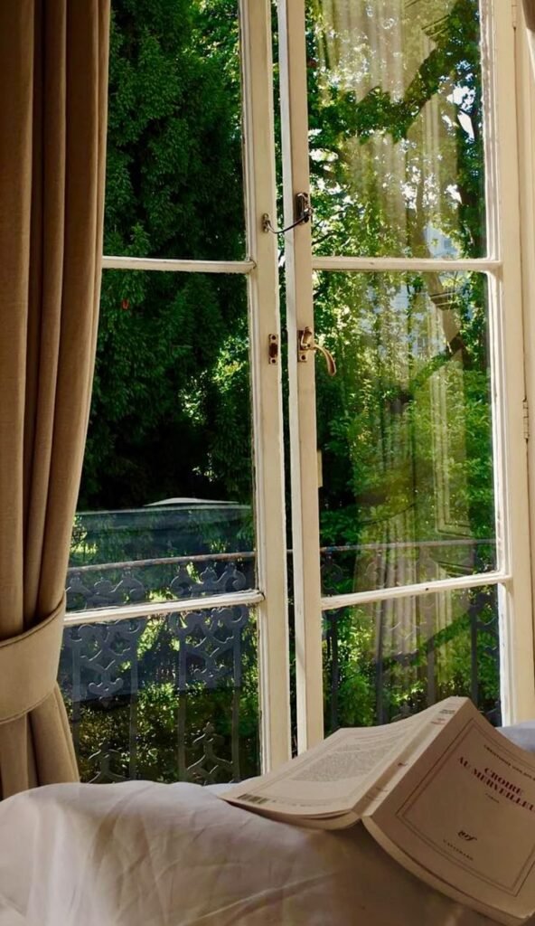 Fenêtre ouverte sur balcon végétalisé avec livre posé pour un moment de lecture estival, à voir dans notre article sur Aménager un coin d’été chez soi sur Deliciously Home