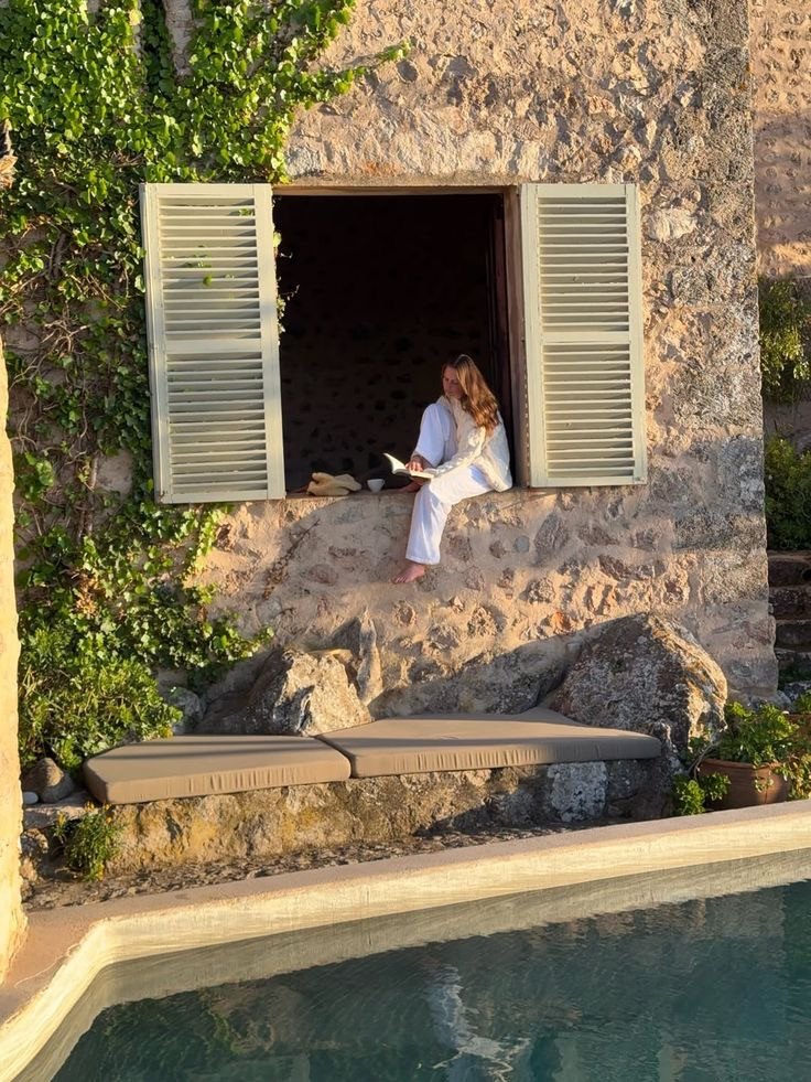 Fenêtre en pierre avec personne lisant et vue sur piscine pour un moment calme en été, à découvrir dans notre article sur Aménager un coin d’été chez soi sur Deliciously Home