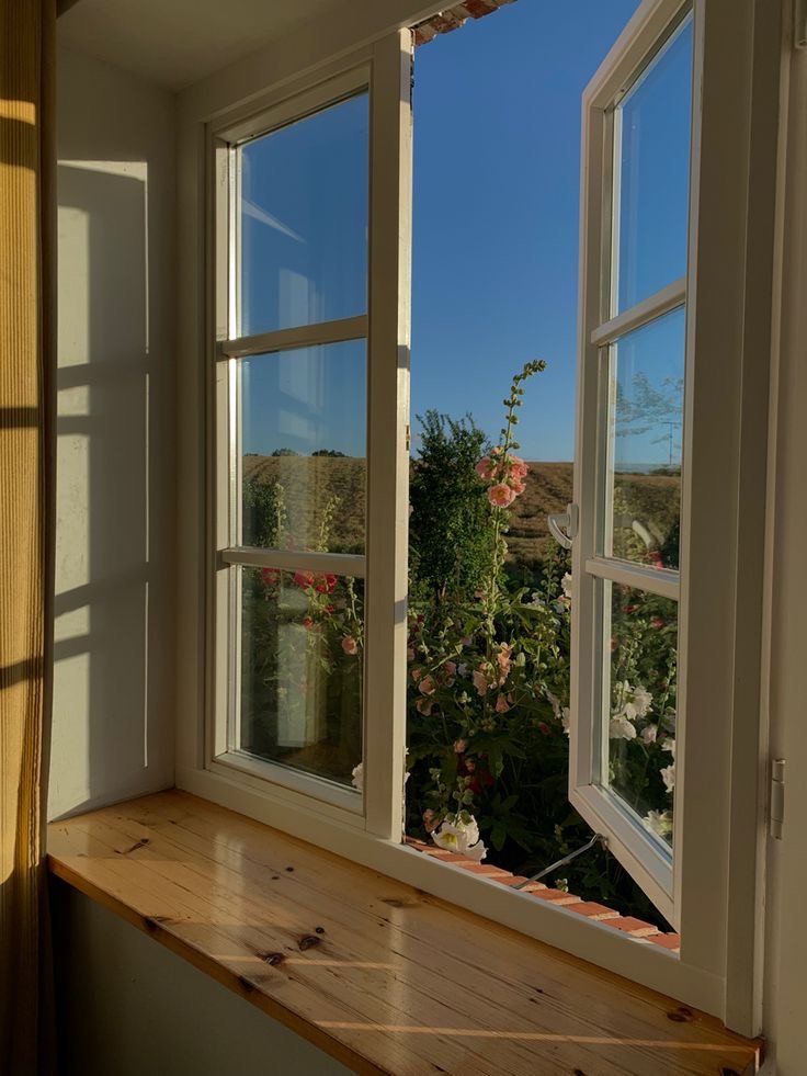 Fenêtre ouverte sur paysage avec fleurs et lumière d’été pour un coin apaisant chez soi, à retrouver dans notre article sur Aménager un coin d’été chez soi sur Deliciously Home