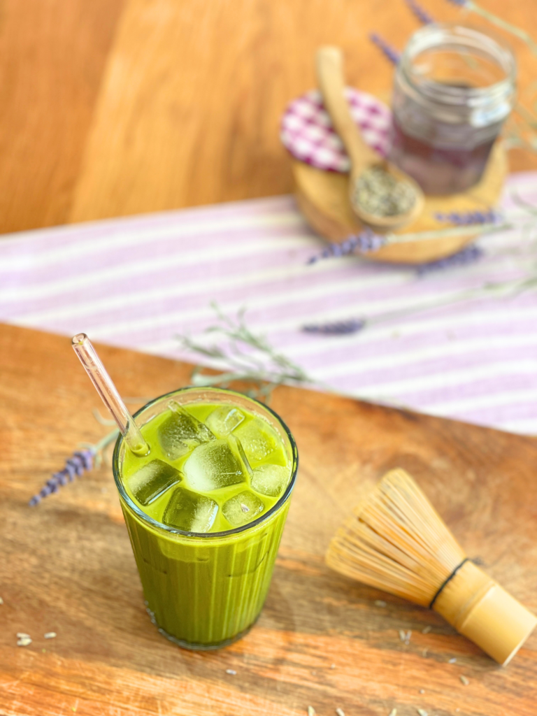 Matcha latte glacé à la lavande servi avec glaçons et paille en verre sur une planche en bois. Une boisson fraîche et crémeuse réalisée par Deliciously Home.