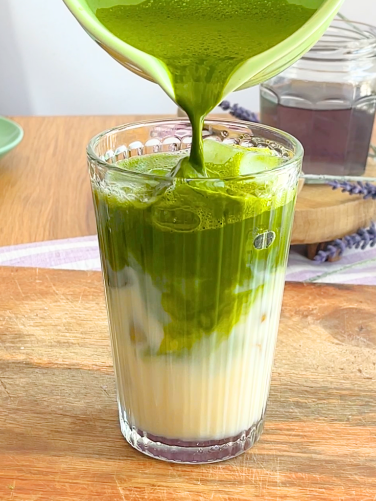 Préparation d’un iced matcha latte lavande avec matcha fouetté versé sur lait végétal et glaçons. Une recette originale et estivale imaginée par Deliciously Home.