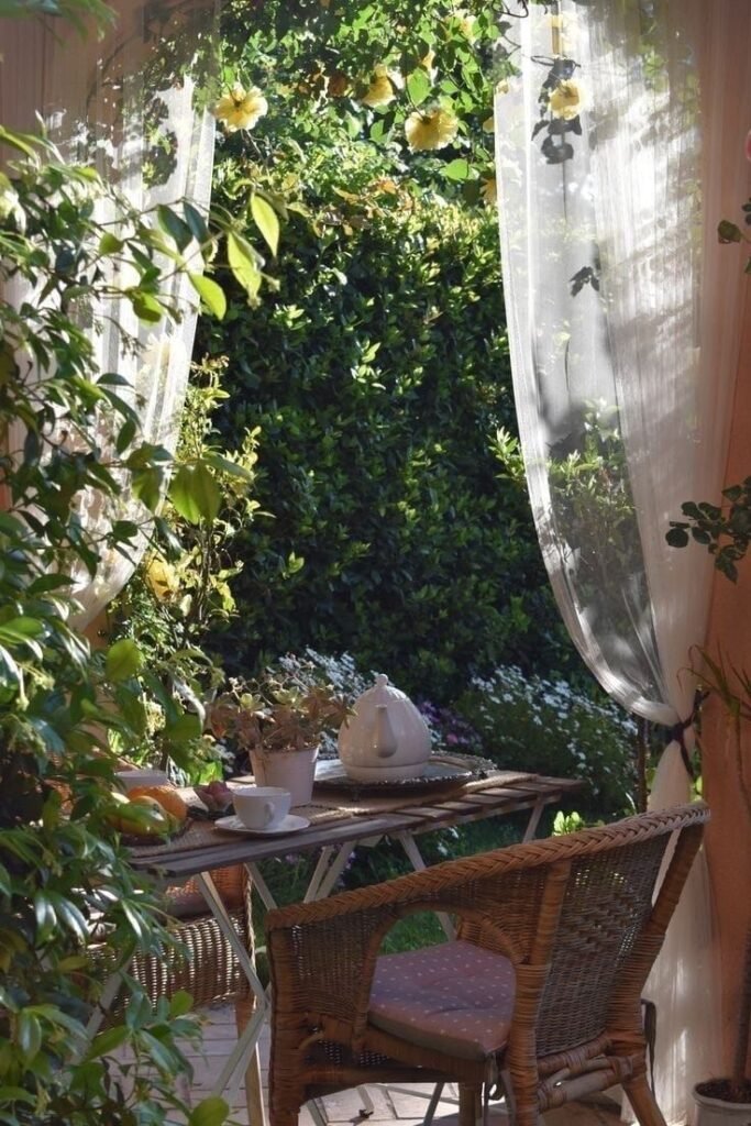 Coin repas extérieur avec table en bois, fauteuil en rotin et rideaux légers entourés de végétation, à explorer dans notre article sur Aménager un coin d’été chez soi sur Deliciously Home
