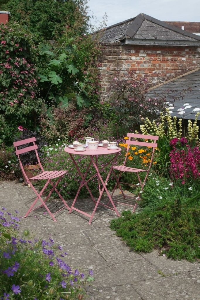 Table et chaises pliantes roses dans un jardin fleuri pour un coin détente plein de charme, à découvrir dans notre article sur Aménager un coin d’été chez soi sur Deliciously Home