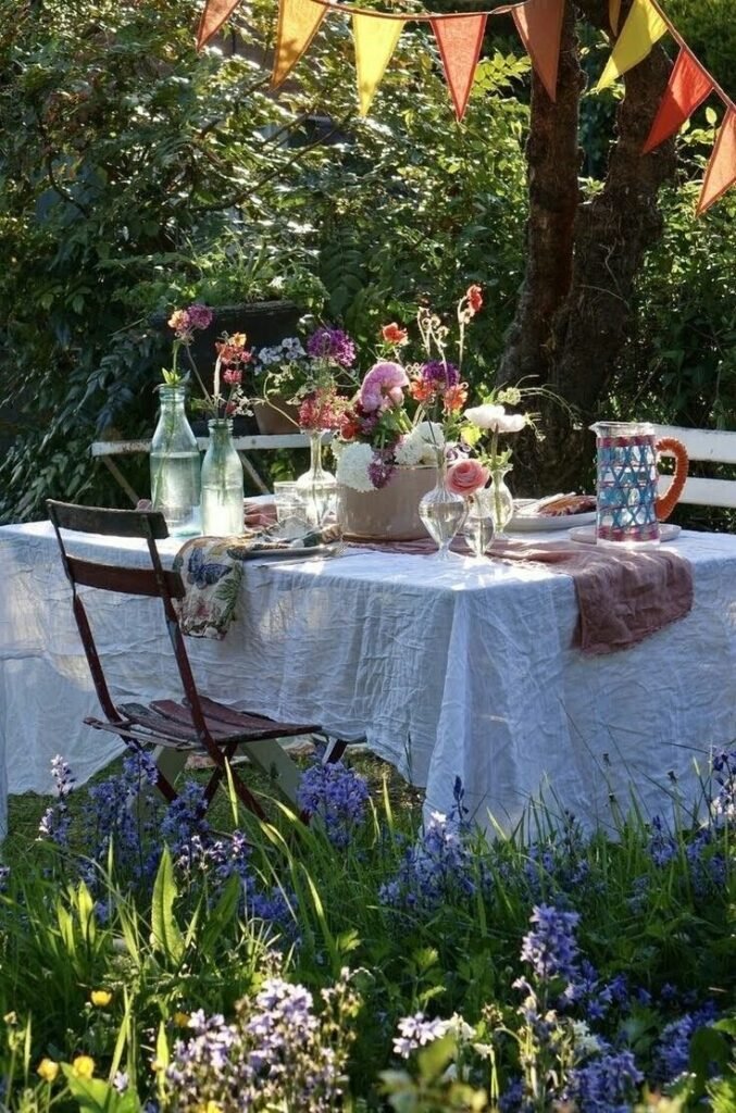 Table dressée en extérieur avec nappe blanche, fleurs et guirlande pour un repas d’été au jardin, à retrouver dans notre article sur Aménager un coin d’été chez soi sur Deliciously Home