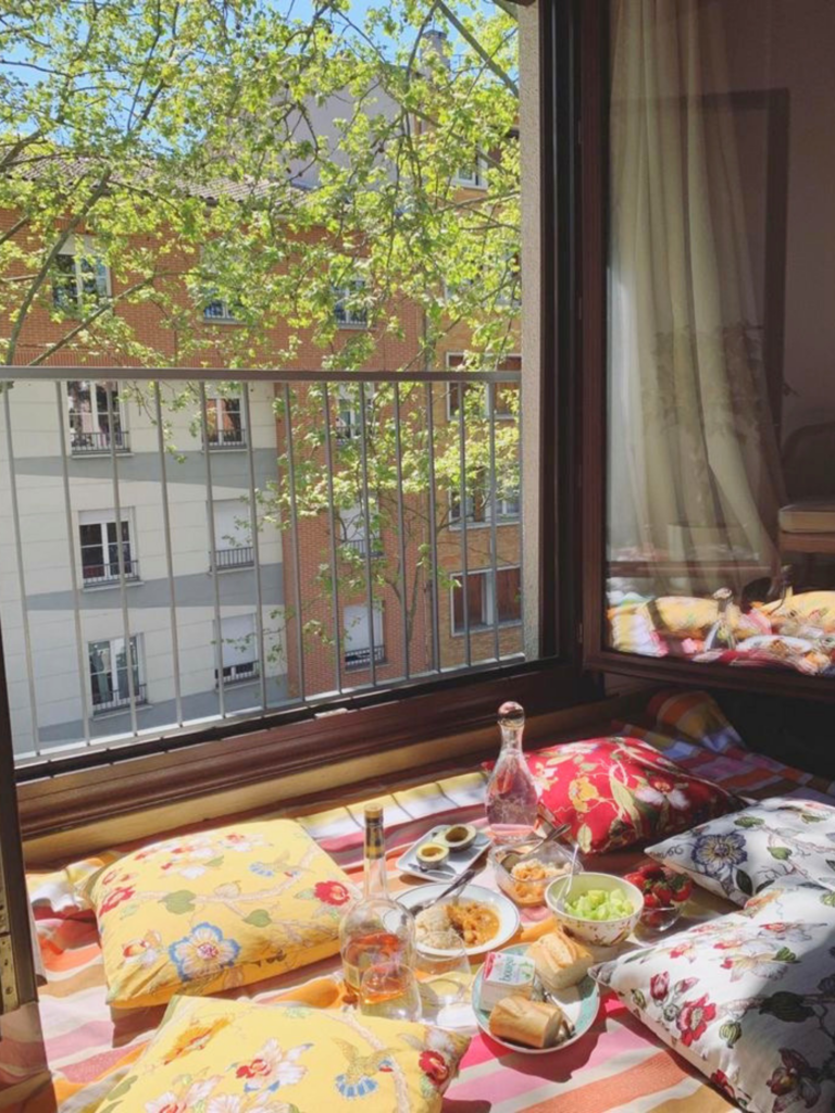 Devant une fenêtre ouverte, coussins, lumière naturelle et assiettes à partager recréent une ambiance pique-nique intime, parfaite pour profiter de l’été autrement. À découvrir avec Deliciously Home
