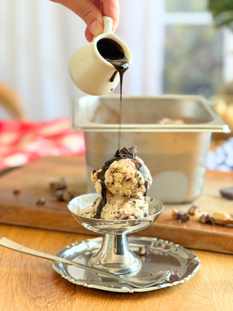 Sauce fudge versée sur une glace Tin Roof maison à la vanille et aux cacahuètes chocolatées. Recette à découvrir sur Deliciously Home.