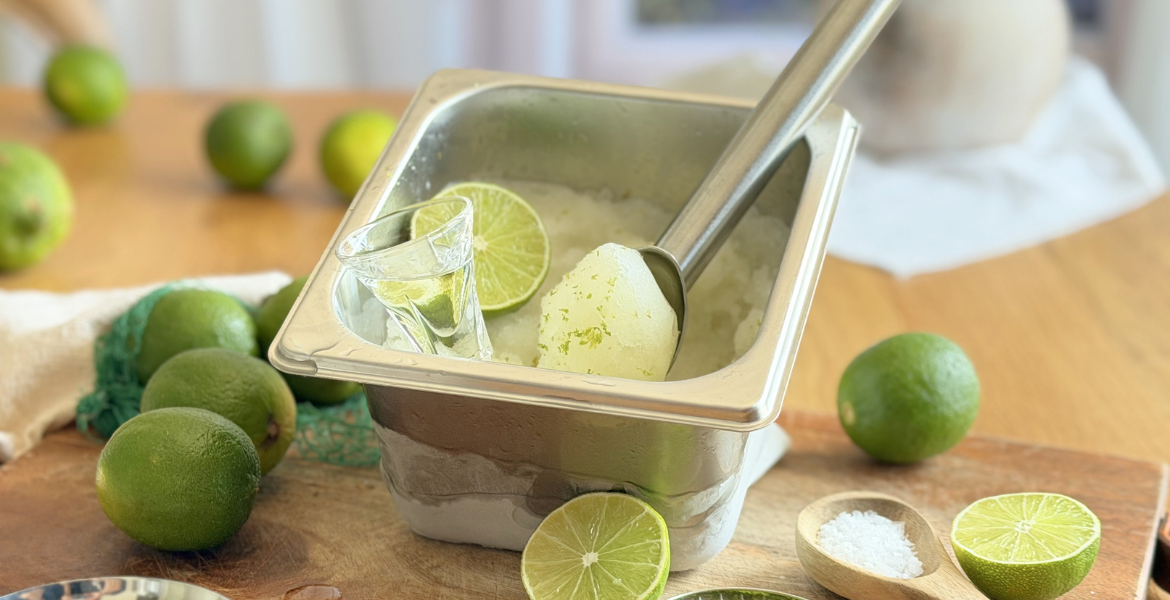 Sorbet au citron vert maison dans un bac inox avec une boule prête à servir, entouré de citrons verts, de fleur de sel et de zestes frais. Découvrez la recette complète en lisant notre article sur Deliciously Home.