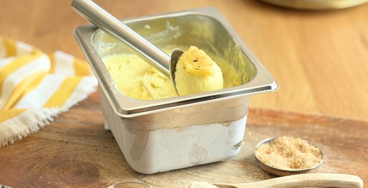 Sorbet Piña Colada maison dans un bac inox avec une boule garnie d'un morceau d'ananas rôti, accompagné de noix de coco râpée. Découvrez la recette complète en lisant notre article sur Deliciously Home.