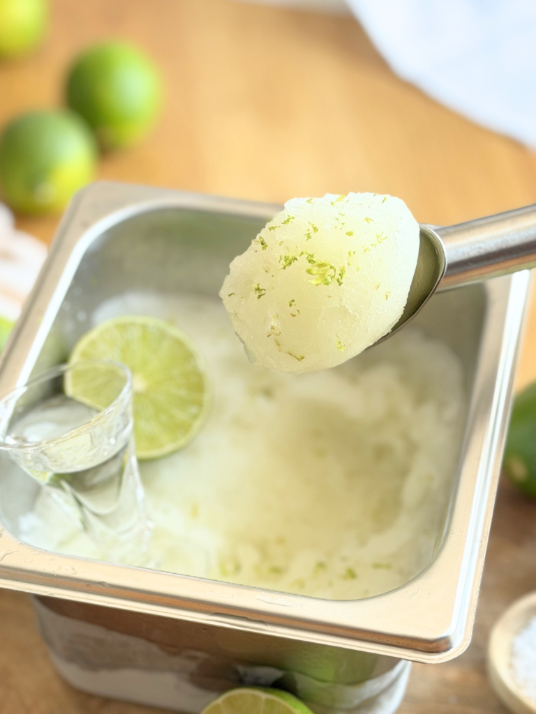 Boule de sorbet au citron vert servie dans une cuillère à glace au-dessus d'un bac inox, avec zestes de citron vert, demi-citron vert et petit verre en arrière-plan. Découvrez la recette complète en lisant notre article sur Deliciously Home.