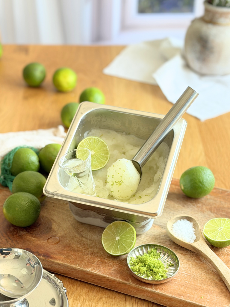 Bac de sorbet au citron vert maison accompagné de citrons verts frais, de fleur de sel, de zestes et d'une cuillère à glace, présenté sur une planche en bois. Découvrez la recette complète en lisant notre article sur Deliciously Home.