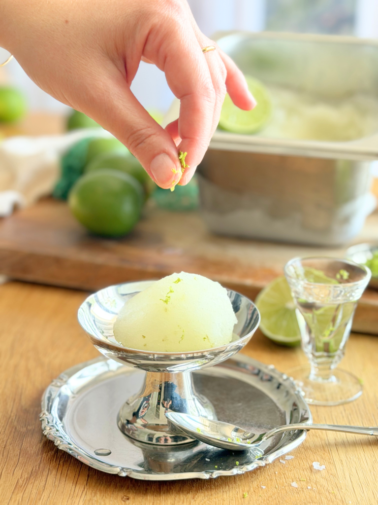 Sorbet au citron vert dressé dans une coupe en métal, parsemé de zestes frais juste avant le service, avec un petit verre et des citrons verts en décoration. Découvrez la recette complète en lisant notre article sur Deliciously Home.
